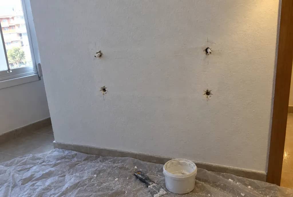 Pared blanca con cuatro agujeros de tacos tras quitar una balda, con una espátula y un bote de masilla sobre un plástico en el suelo
