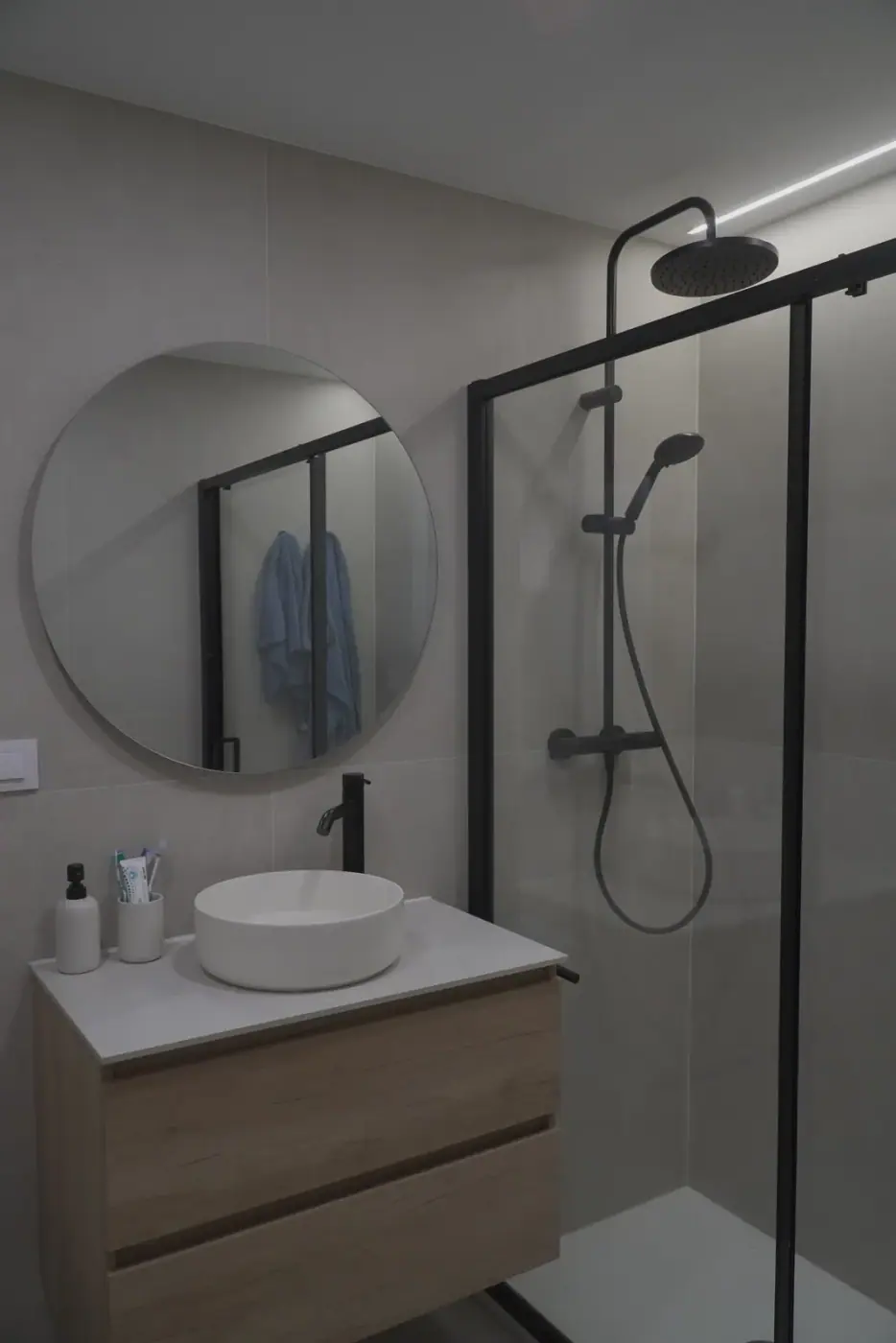 Vista completa de baño reformado con ducha de perfil negro, mueble de madera y espejo redondo