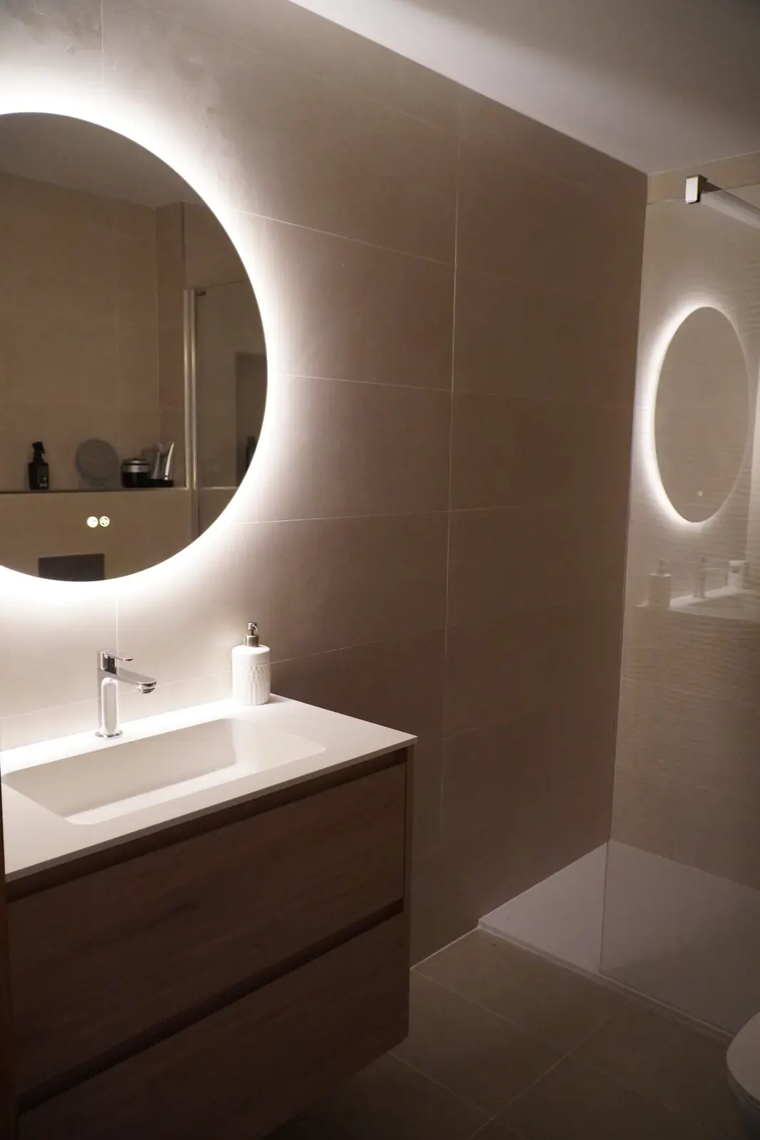 Baño reformado con espejo redondo retroiluminado y microcemento beige