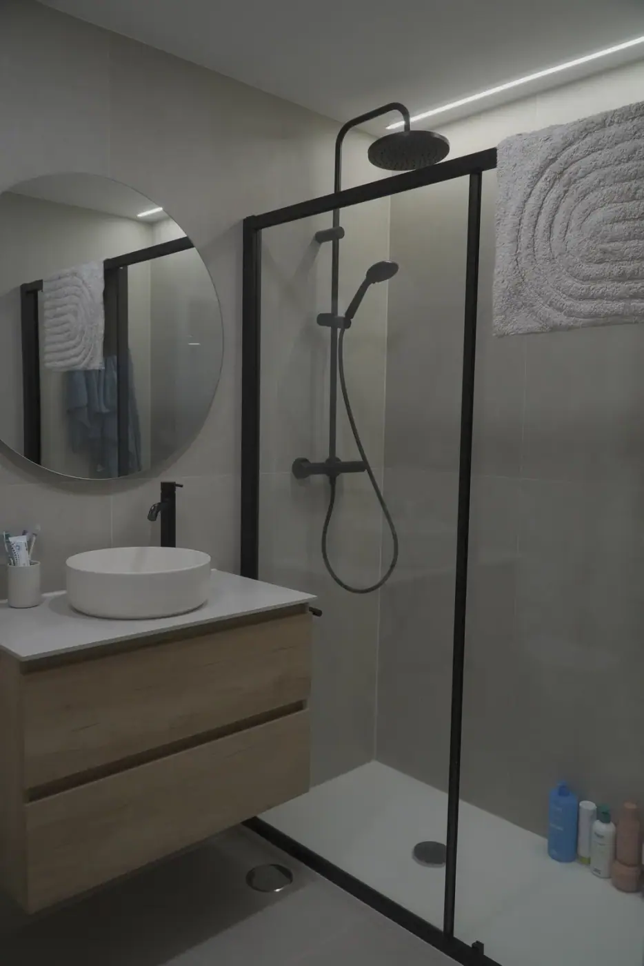 Baño reformado con mampara de perfil negro y espejo redondo sobre el mueble