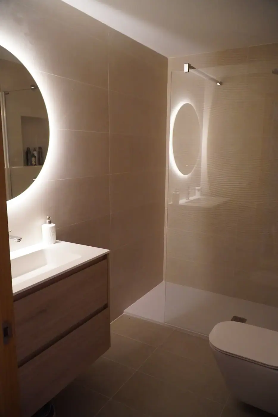 Espejo redondo con luz indirecta reflejando la ducha del baño reformado