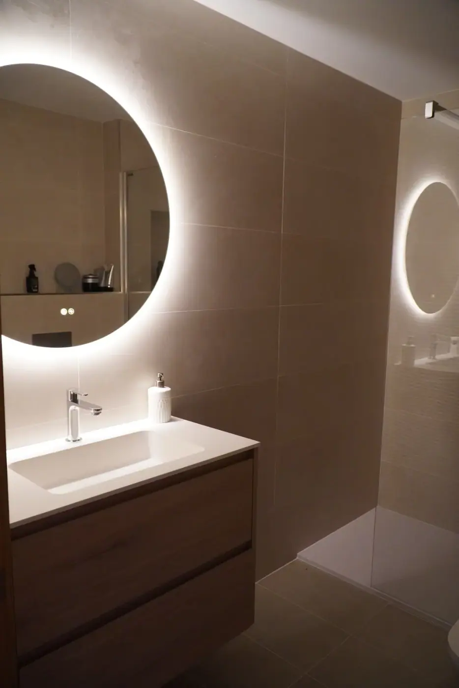 Lavabo sobre encimera y espejo redondo retroiluminado en baño de tonos arena